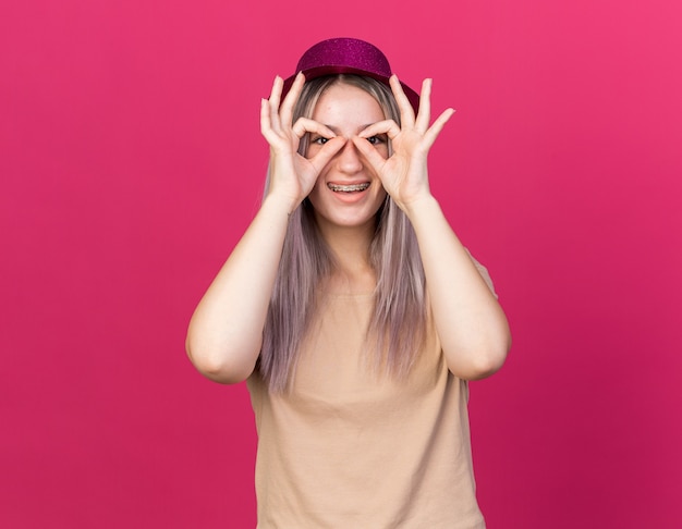 Smiling young beautiful girl wearing party hat with dental braces showing look gesture isolated on pink wall