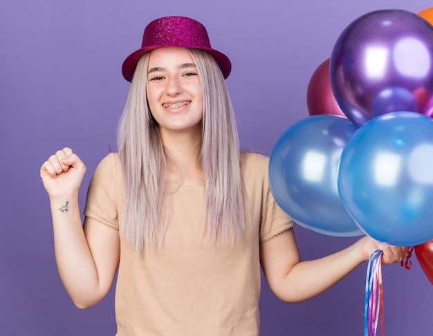 Smiling young beautiful woman wearing dental braces and party hat holding balloons showing yes gesture isolated on blue wall