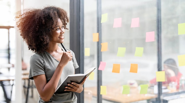 Smiling young black businesswoman holding notebook looking and thinking standing next to a glass wall