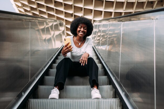 Smiling young black teenage woman with afro hair sitting on an outdoor escalator taking a selfie