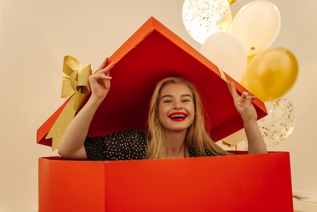 Smiling young blonde caucasian girl sits in big red box making surprise to her boyfriend Holiday concept