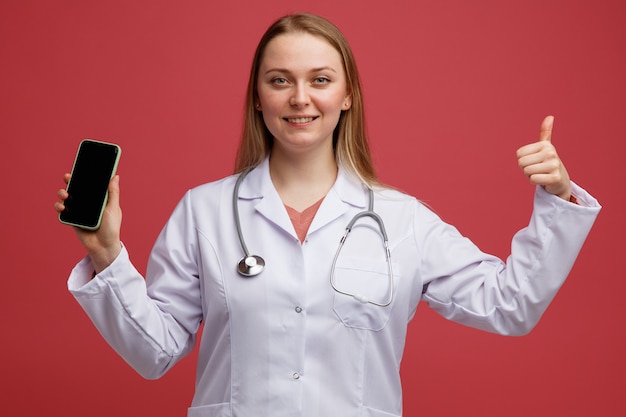 Smiling young blonde female doctor wearing medical robe and stethoscope around neck holding mobile phone showing thumb up 