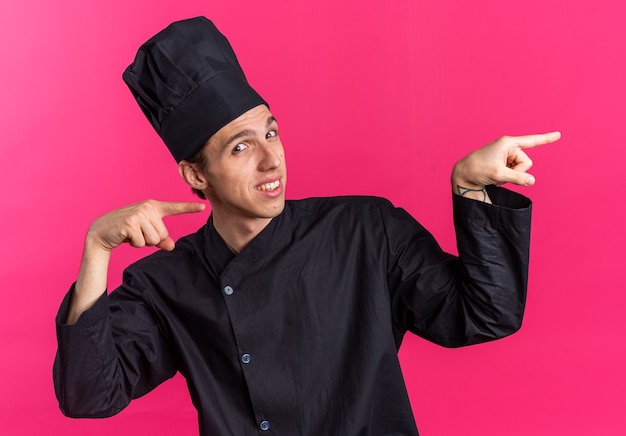 Smiling young blonde male cook in chef uniform and cap looking at camera pointing at side isolated on pink wall