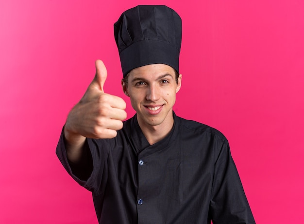 Smiling young blonde male cook in chef uniform and cap looking at camera showing thumb up isolated on pink wall