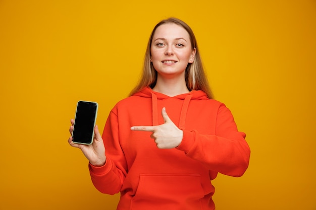 Smiling young blonde woman holding and pointing at mobile phone 