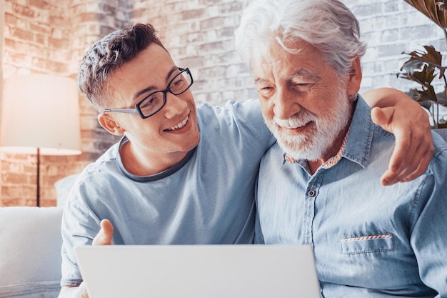 Smiling young boy helping senior grandfather browse with laptop and choose goods or services on web surfing together at home Younger generation caring about older relatives teaching using computer