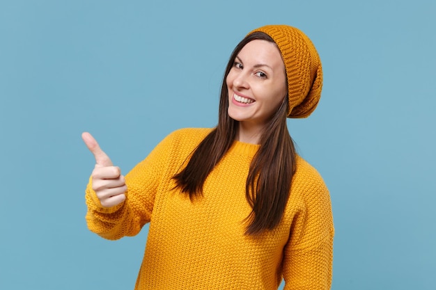 Smiling young brunette woman girl in yellow sweater and hat posing isolated on blue wall background studio portrait. People sincere emotions lifestyle concept. Mock up copy space. Showing thumb up.