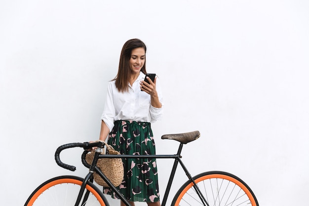 Smiling young brunette woman wearing summer clothes standing at the wall outdoors, holding bicycle, using mobile phone