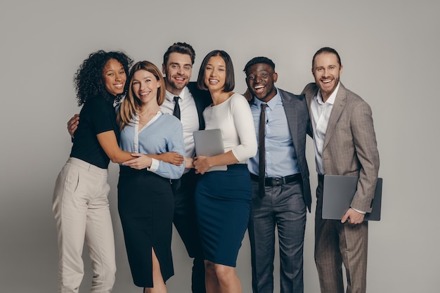 Smiling young business team in formalwear embracing while standing on beige background