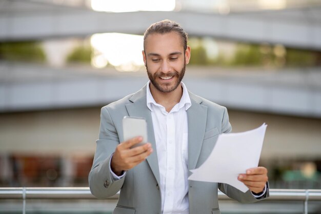 Smiling young businessman comparing document with information on his smartphone