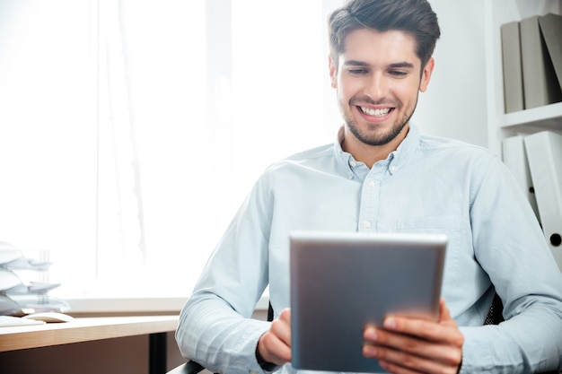 Smiling young businessman sitting at the table and using tablet computer in office