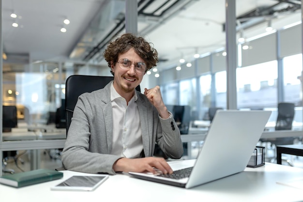 Smiling young businessman with curly hair focused on work at his laptop in a welllit modern