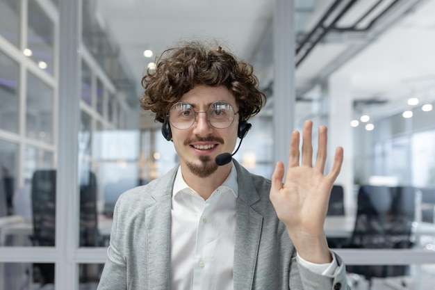 Smiling young businessman with curly hair and headset waving in office representing a friendly