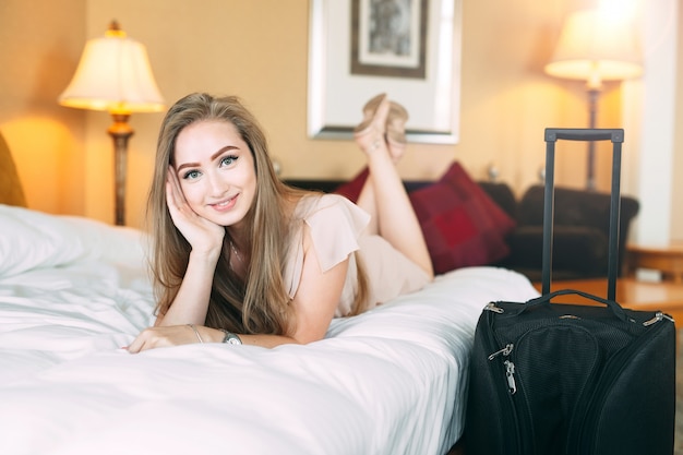 Smiling young businesswoman in bed, girl licks on the bed near the suitcase.