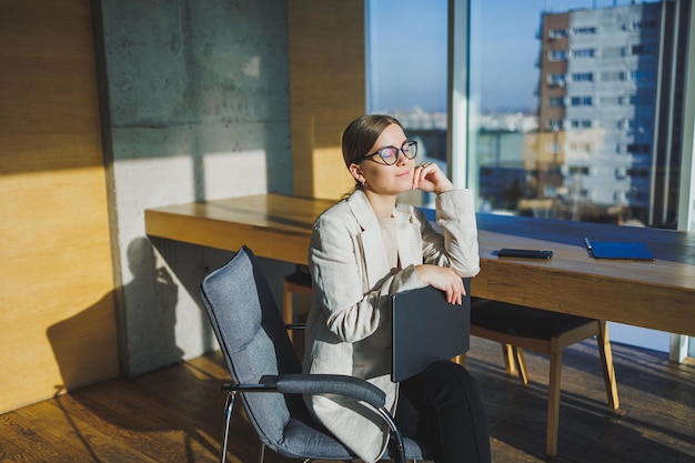 Smiling young businesswoman in casual clothes and glasses sitting in the office with a mobile phone while working on a remote project using a laptop