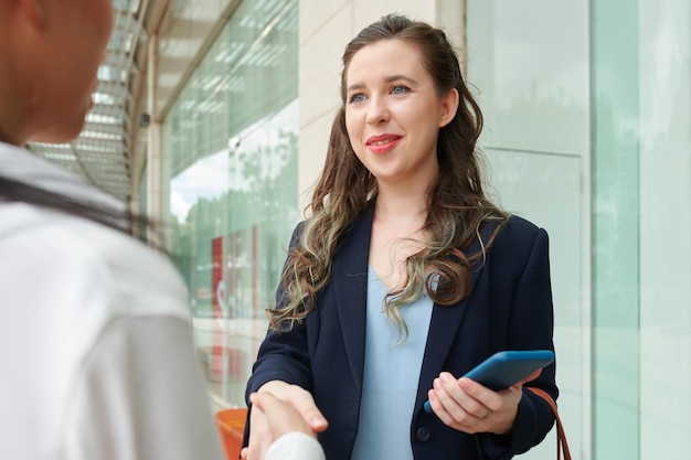 Smiling young businesswoman with smartphone shaking hand of business partner