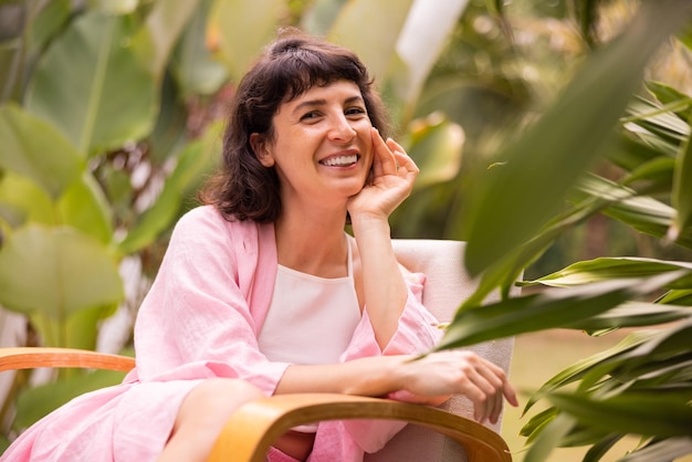 Smiling young caucasian brunette girl in pink clothes looks at camera sitting on chair among nature in garden Vacation happiness and pleasure concept