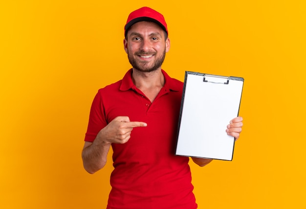 Smiling young caucasian delivery man in red uniform and cap looking at camera showing clipboard pointing at it isolated on orange wall