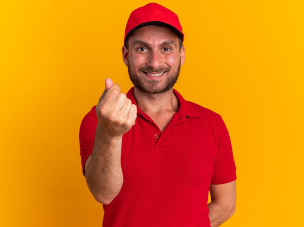 Smiling young caucasian delivery man in uniform and cap keeping hand behind back doing money gesture 