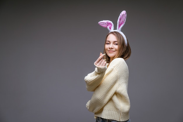 smiling young caucasian girl, blonde with bunny ears, shows a mini heart with fingers isolated over gray wall