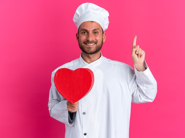 Smiling young caucasian male cook in chef uniform and cap holding heart shape pointing up 