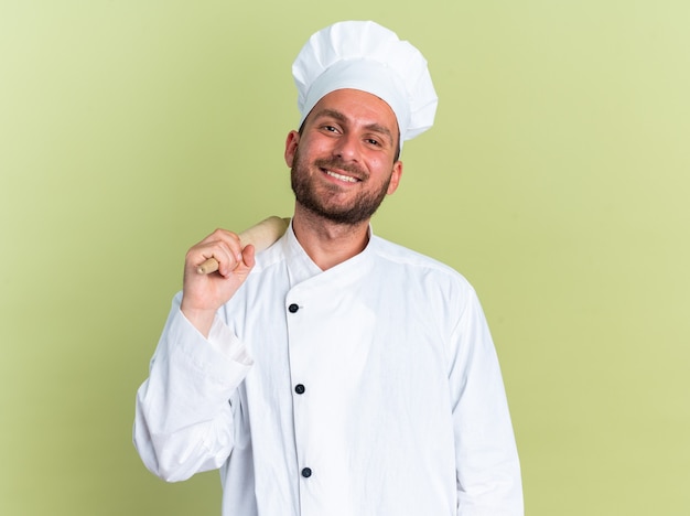 Smiling young caucasian male cook in chef uniform and cap holding rolling pin on shoulder 