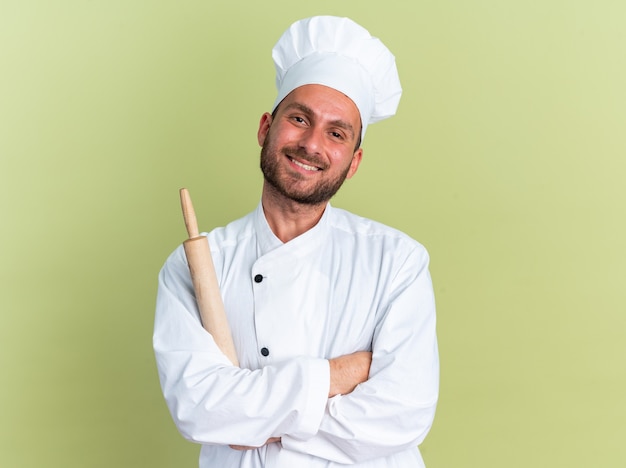 Smiling young caucasian male cook in chef uniform and cap standing with closed posture holding rolling pin looking at camera isolated on olive green wall with copy space