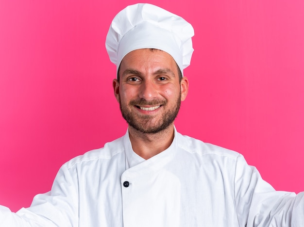 Smiling young caucasian male cook in chef uniform and cap stretching out hands looking at camera isolated on pink wall