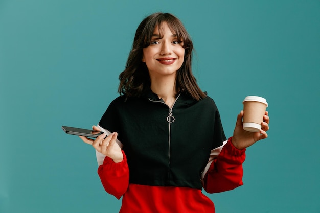 Smiling young caucasian woman holding mobile phone and paper coffee cup looking at camera isolated on blue background