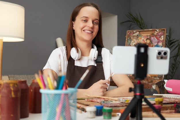 Smiling young Caucasian woman wearing brown apron painting with brush in artist studio using smartphone and tripod recording tutoring video to her internet channel