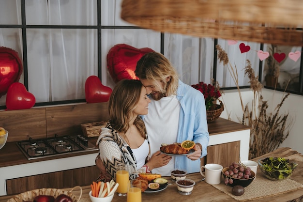 Smiling young couple having breakfast at the kitchen with red heart shape balloons on background