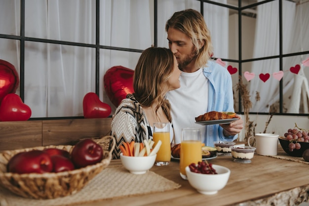 Smiling young couple kissing while having breakfast at the kitchen with red balloons on background