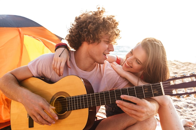 Smiling young couple resting together at the beach, camping, playing guitar