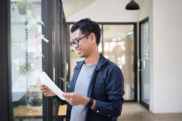 Smiling young creative designer reading document while standing near glass wall in modern office