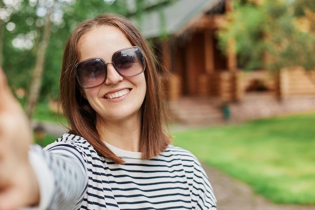 Smiling young dark haired girl making selfie photo in park with wooden house on background expressing positive emotions POV woman broadcasting livestream having video call