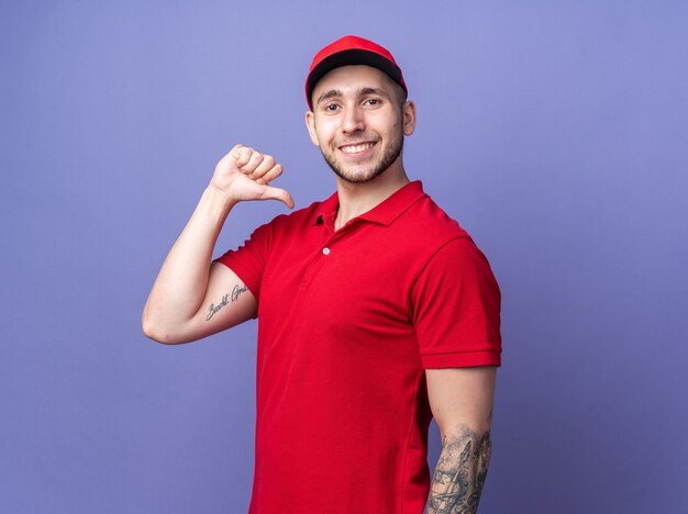 Smiling young delivery guy wearing uniform with cap points at himself 