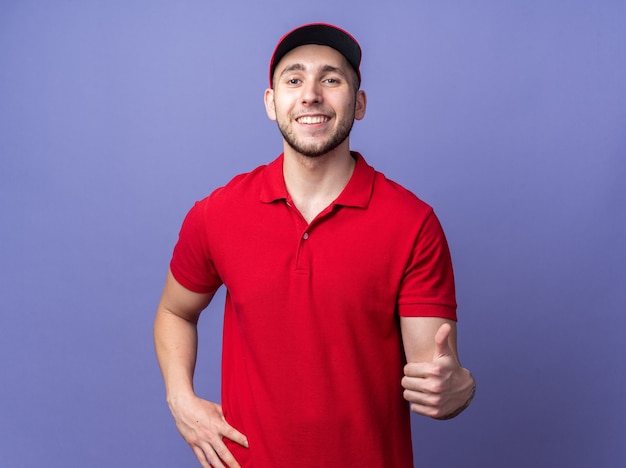 Smiling young delivery guy wearing uniform with cap showing thumb up putting hand on hip 