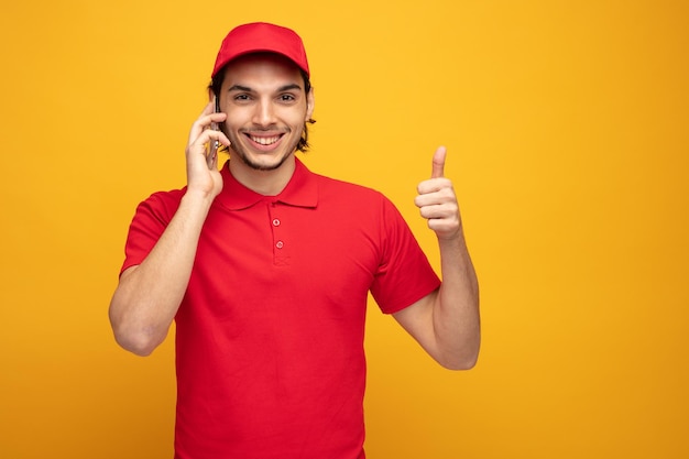 smiling young delivery man wearing uniform and cap looking at camera showing thumb up while talking on phone isolated on yellow background