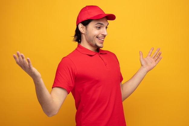 smiling young delivery man wearing uniform and cap standing in profile view looking at side showing empty hands isolated on yellow background