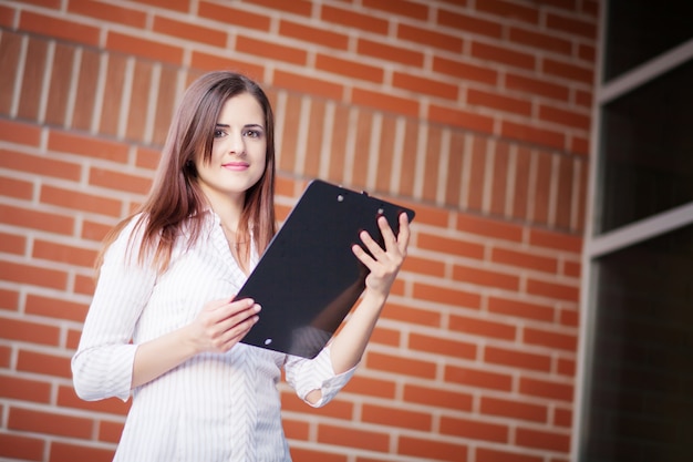 Smiling young elegant businesswoman with clipboard standing in a bright office 