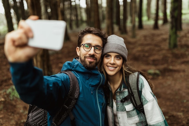 Smiling young european couple tourists in jackets with backpack walk in autumn forest take selfie