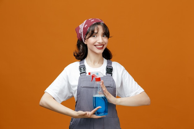 Smiling young female cleaner wearing uniform and bandana holding cleanser with both hands looking at camera isolated on orange background