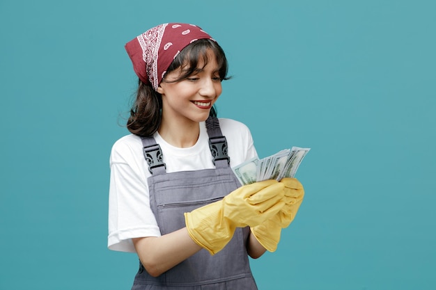 Smiling young female cleaner wearing uniform bandana and rubber gloves counting money biting lip isolated on blue background