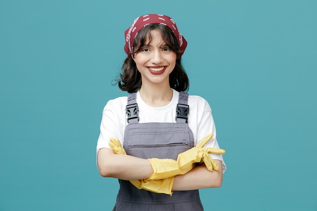 Smiling young female cleaner wearing uniform bandana and rubber gloves keeping arms crossed looking at camera isolated on blue background