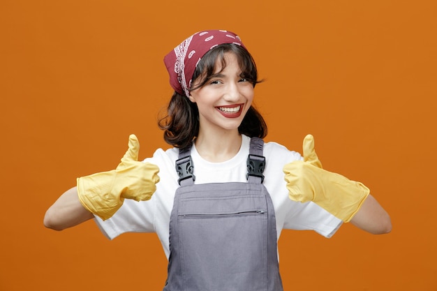 Smiling young female cleaner wearing uniform rubber gloves and bandana looking at camera showing thumbs up isolated on orange background