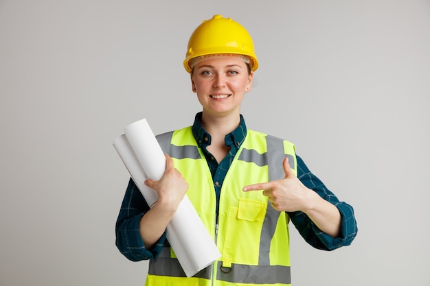 Smiling young female construction worker wearing safety helmet and safety vest holding and pointing at papers 