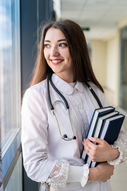 Smiling young female doctor in uniform with stethoscope holding book while standing