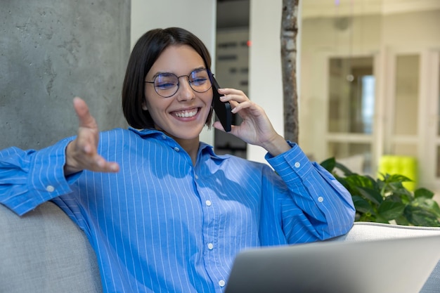 Smiling young female freelancer with the laptop seated on the sofa talking on the cellphone