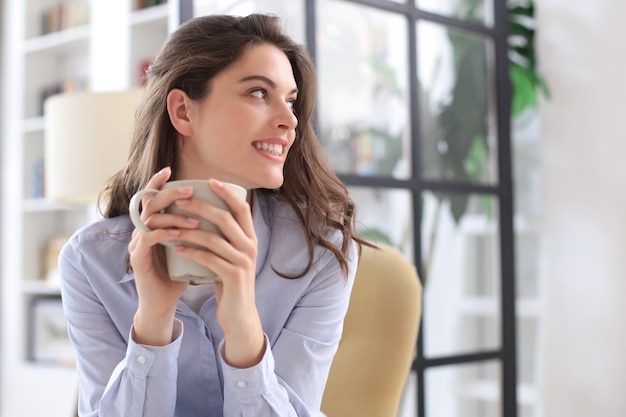 Smiling young female sitting in the armchair in the living room, holding a cup of coffee.