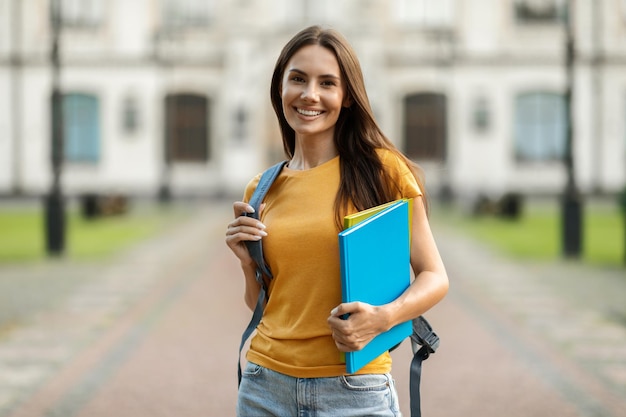 Smiling young female student with workbooks and backpack standing outdoors